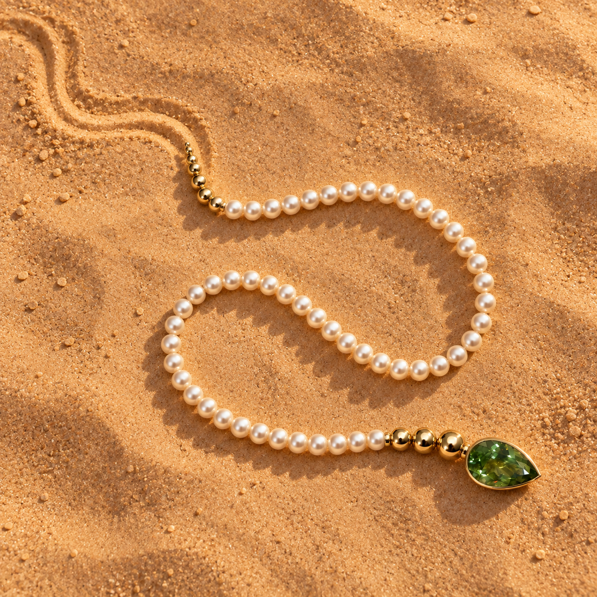 Pearl necklace with gold beads and a pear-shaped green gemstone clasp, arranged in an S-shaped curve on golden sand.