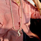 Alphabetty gold S pendant on a long pearl necklace over a red-and-white gingham shirt, layered with a gold dog necklace.