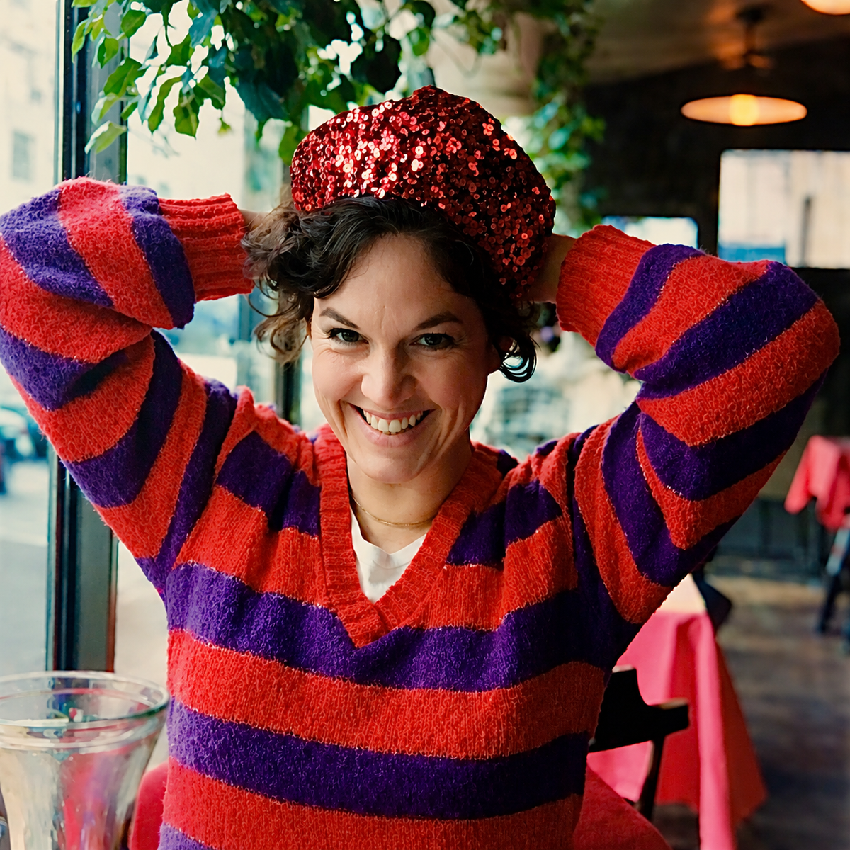 A smiling woman in a red-and-purple striped sweater adjusts a red sequined hat in a café.
