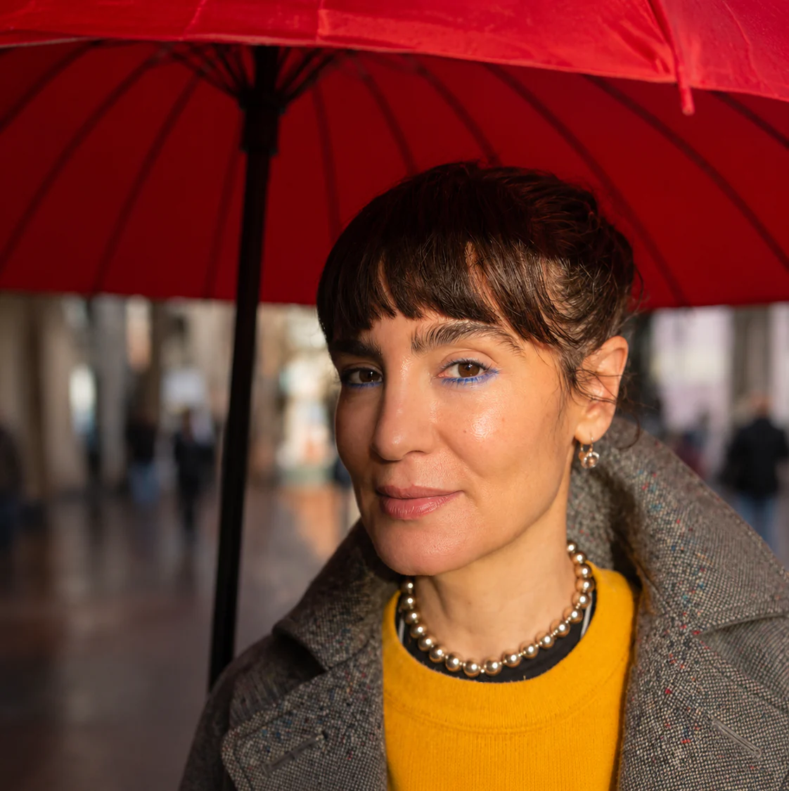 Elisabeth Marques founder Shereen Safadi-Freeman beneath a red umbrella, wearing a gold bead necklace and yellow sweater.