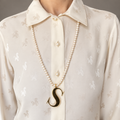 Alphabetty gold letter S pendant worn on a long pearl necklace over a cream blouse.