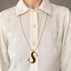 Alphabetty gold letter S pendant worn on a long pearl necklace over a cream blouse.