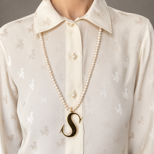 Alphabetty gold letter S pendant worn on a long pearl necklace over a cream blouse.
