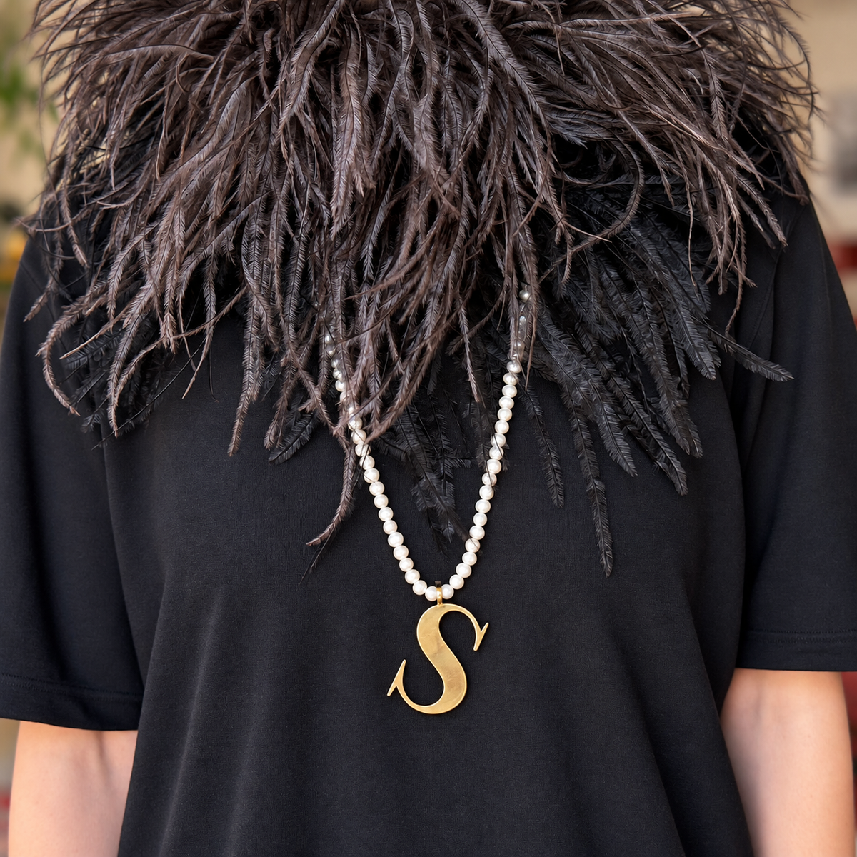 Alphabetty gold S pendant on a long pearl necklace, styled over a black top with a feathered collar.