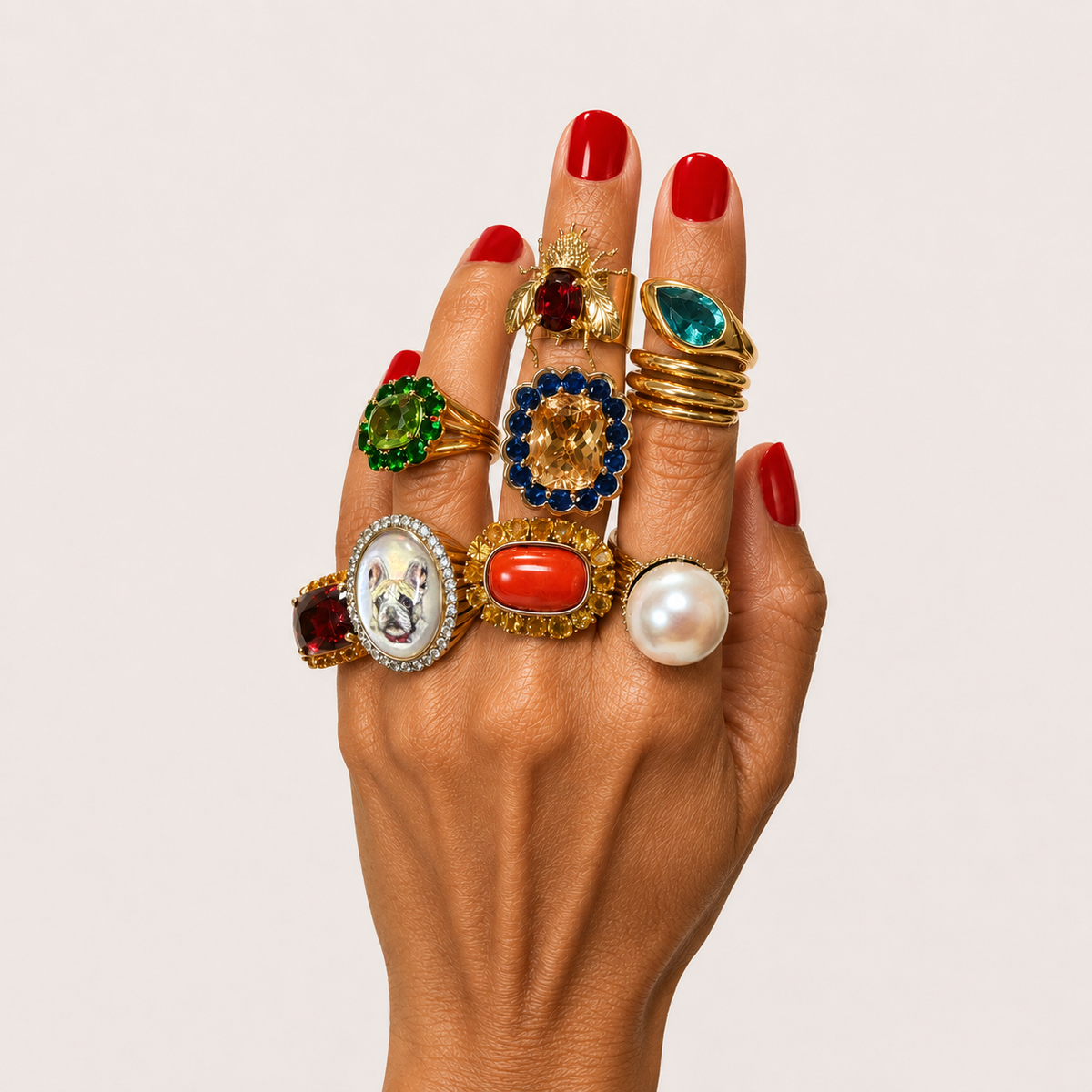 A hand wearing eight Elisabeth Marques statement rings in gold, with colourful gemstones, a pearl, a bumblebee and a pet portrait.