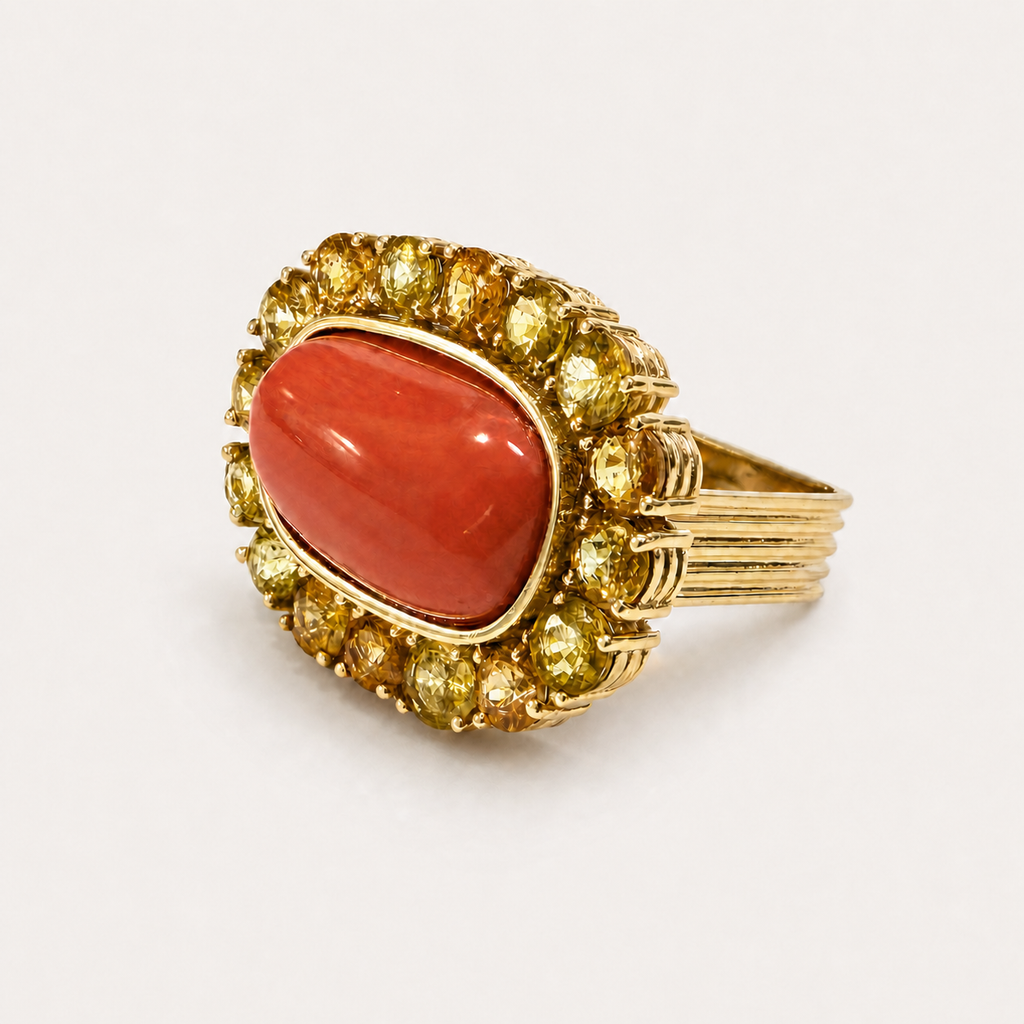 Angled view of the Coralie ring, showing its red Italian coral cabochon, golden sapphire halo and fluted 18K gold shoulders.