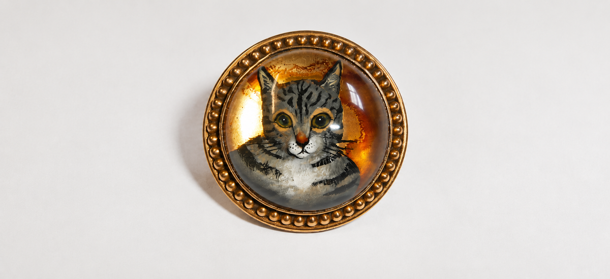 A round decorative brooch featuring a painted tabby cat portrait under a clear dome, surrounded by an ornate gold-toned beaded border, photographed on a plain light background.