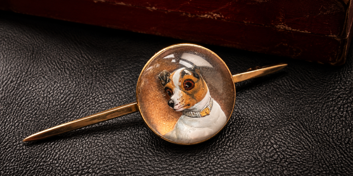 A vintage-style gold-toned lapel pin featuring a circular glass dome with a hand-painted portrait of a small dog wearing a hat and collar, displayed on a dark textured surface.