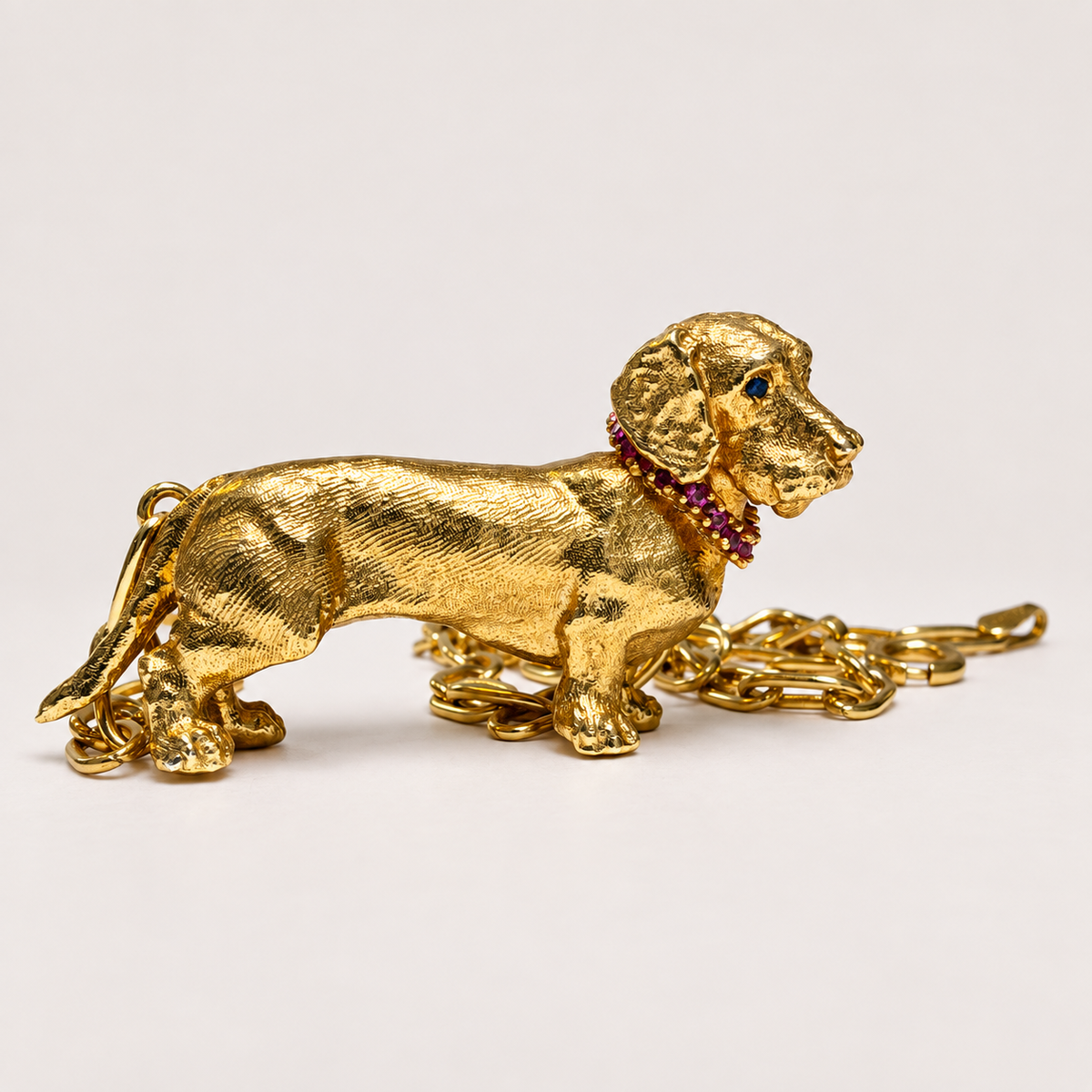 Side view of the Gatsby 18K gold dachshund pendant with a ruby collar and gold chain resting behind it.