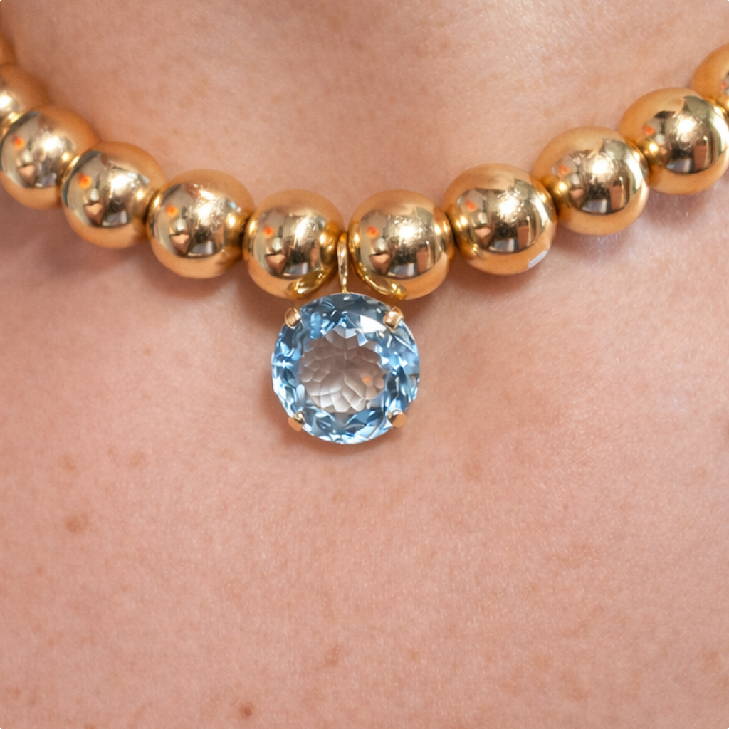 Round blue topaz charm worn at the centre of a necklace of polished gold beads, close-up.