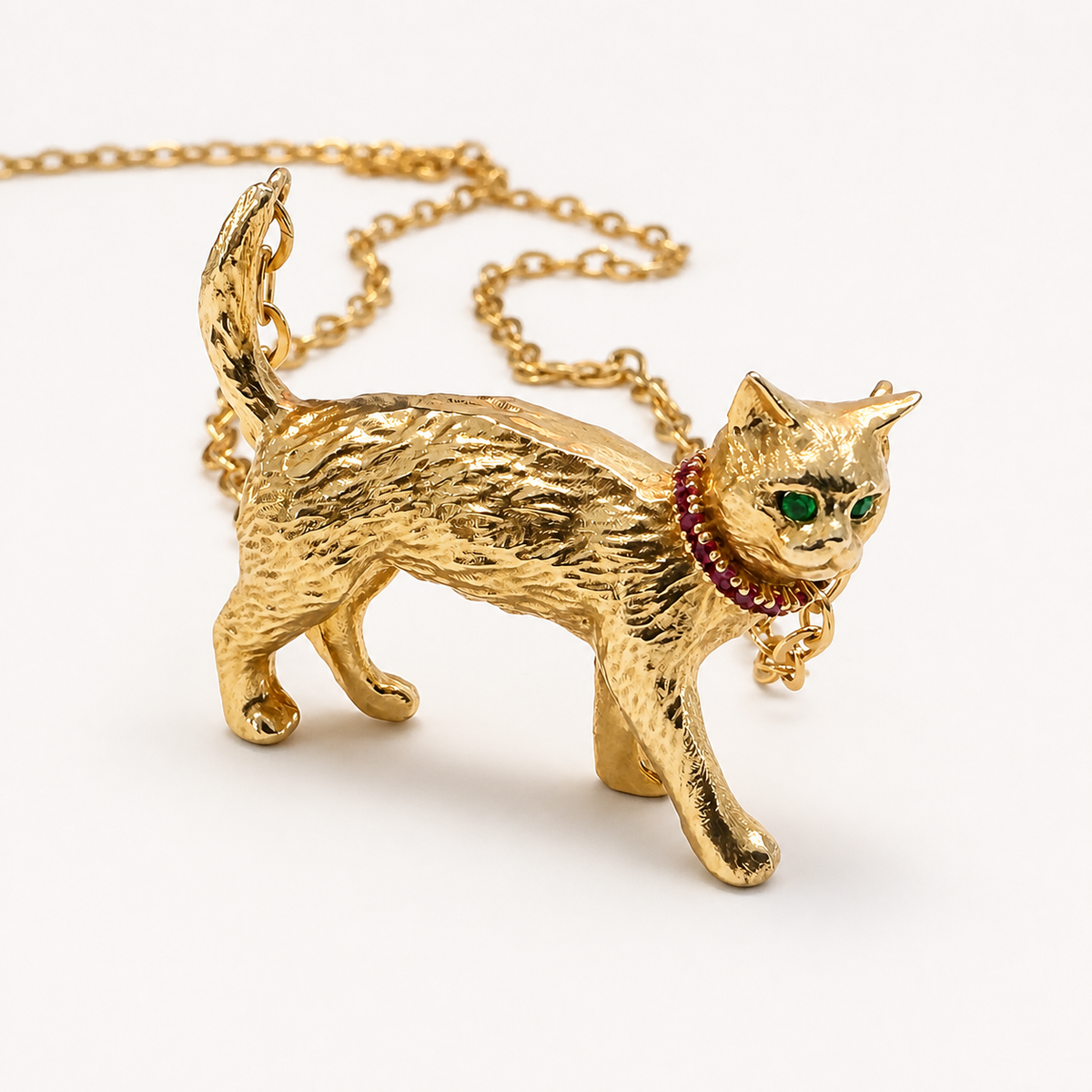 Angled front view of the Gigi gold cat pendant with textured fur, emerald eyes and a ruby collar, with its gold chain behind.