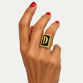 18K gold initial D ring with black onyx and a golden sapphire halo, worn on a hand with red nails.