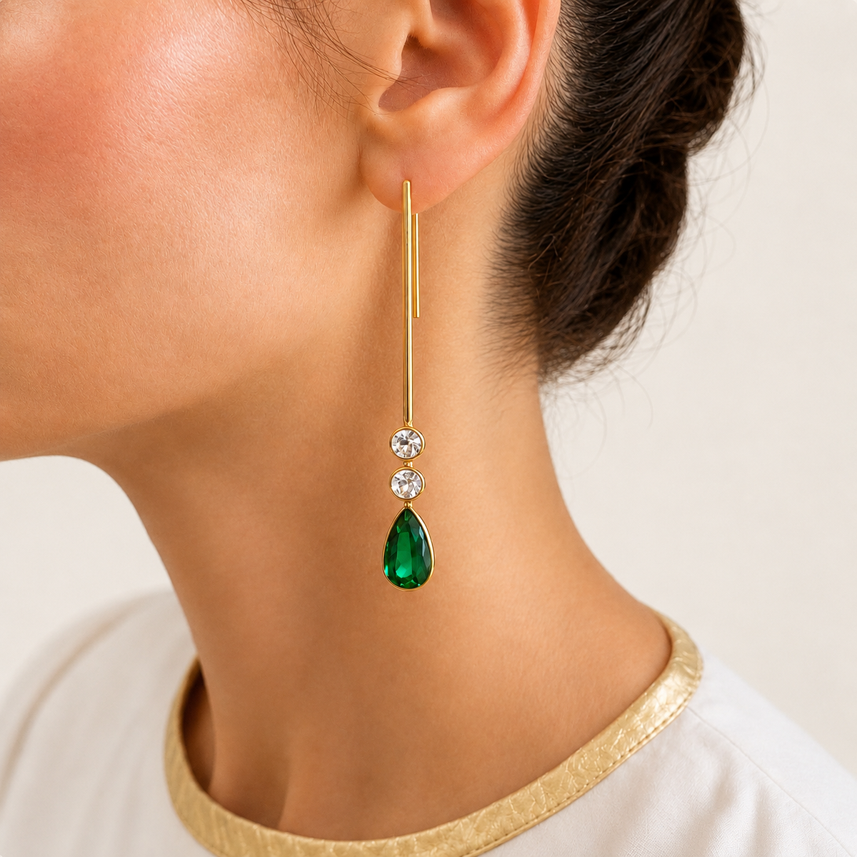 Serena stick earring worn on a model, with a slender gold bar, two round aquamarines and a pear-shaped green emerald drop.