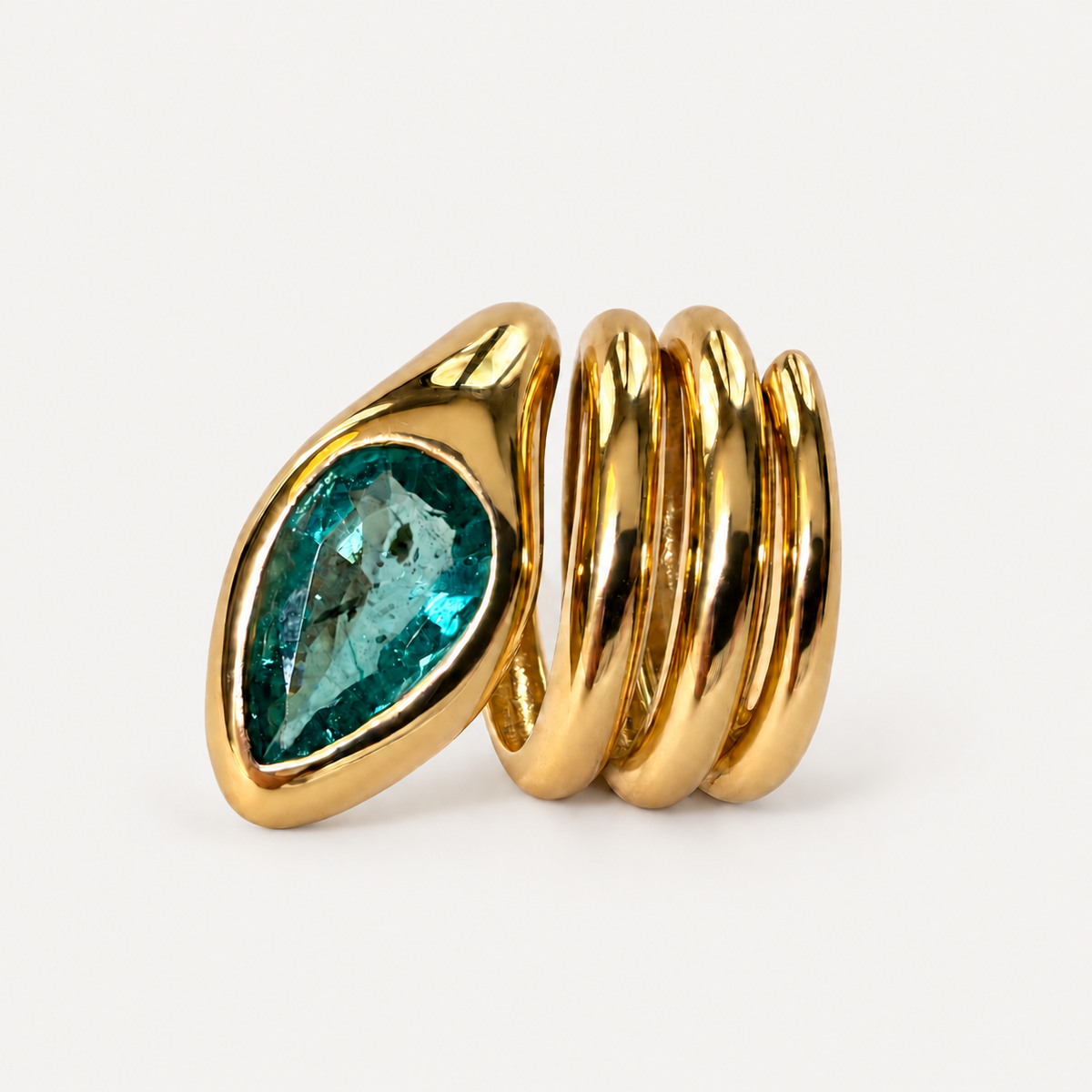 Close-up of the Sylvie ring with its pear-shaped emerald facing forward beside three polished 18K gold coils.